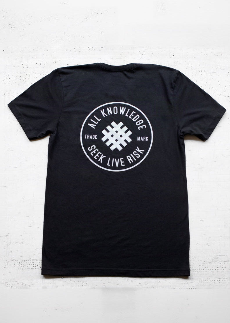 A black short-sleeve t-shirt laid flat on a white textured background, featuring a white circular graphic on the back with the text 'ALL KNOWLEDGE SEEK LIVE RISK' and a geometric symbol in the center.