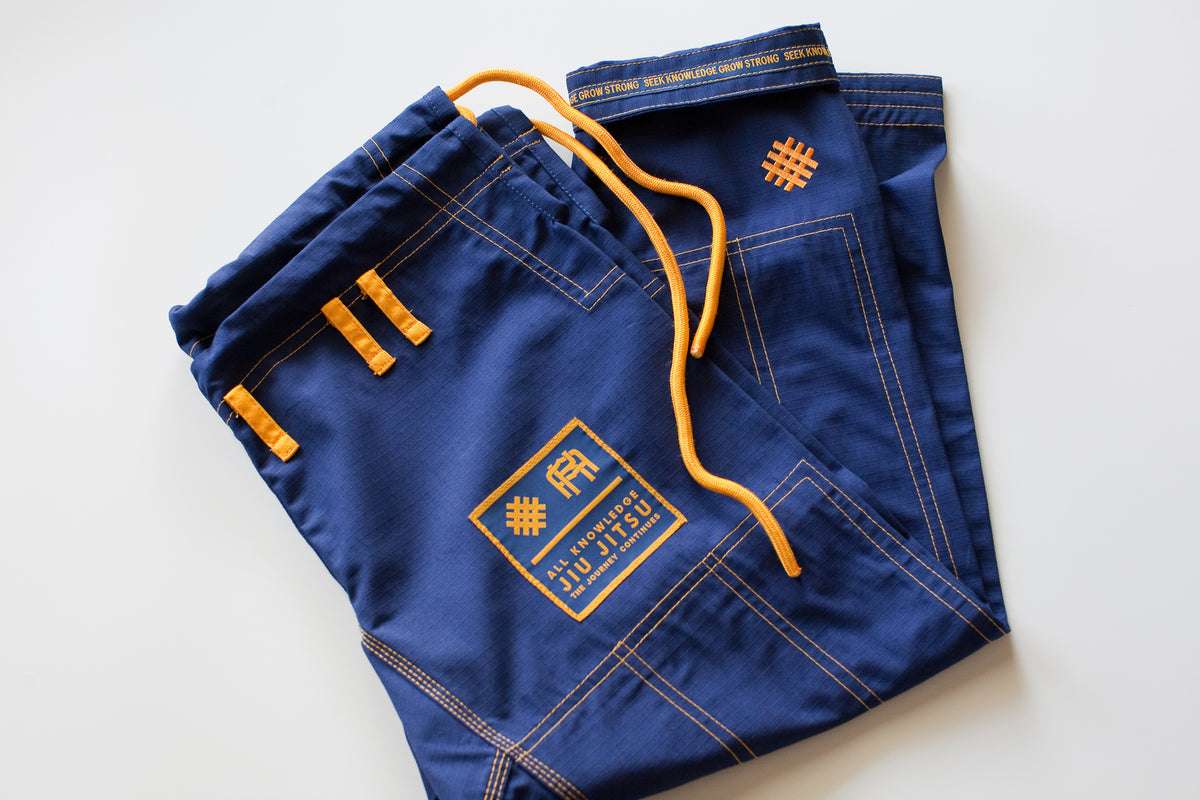 All Knowledge blue Brazilian Jiu-Jitsu Gi with gold stitching, embroidered AK logos, Seek Knowledge Grow Strong sleeve design, matching pants, and branded patches, displayed flat on a white background.