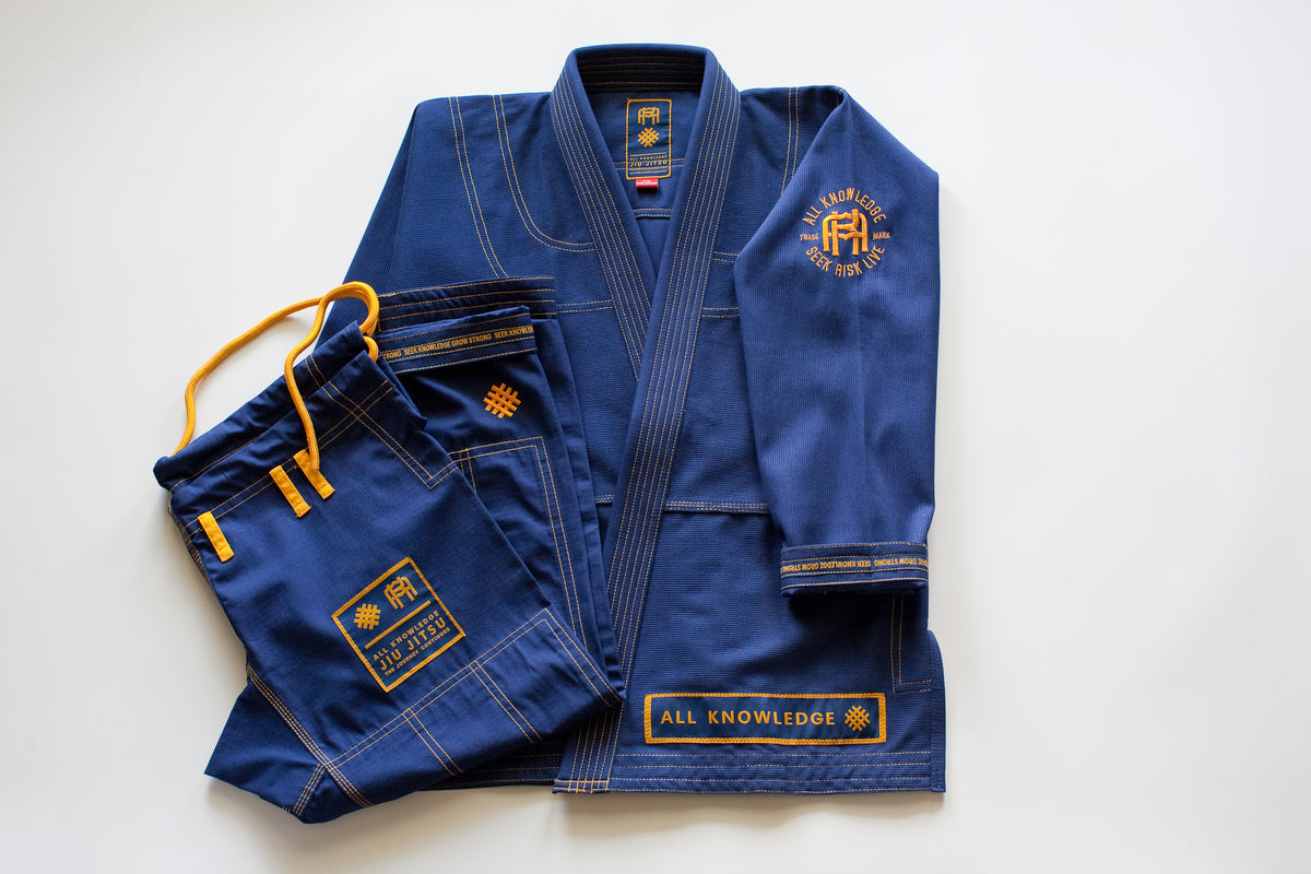 All Knowledge blue Brazilian Jiu-Jitsu Gi with gold stitching, embroidered AK logos, Seek Risk Live sleeve design, matching pants, and branded patches, displayed flat on a white background.